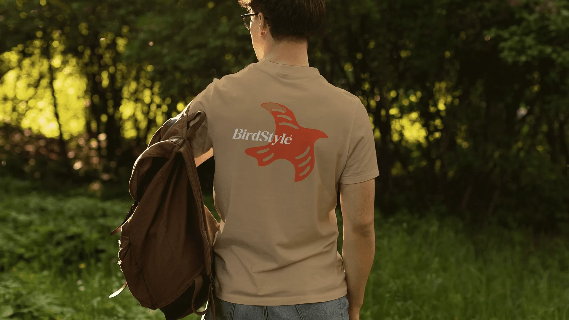Pessoa de costas em trilha na mata usando camiseta bege com estampa grande de pássaro em laranja e o logotipo BirdStyle