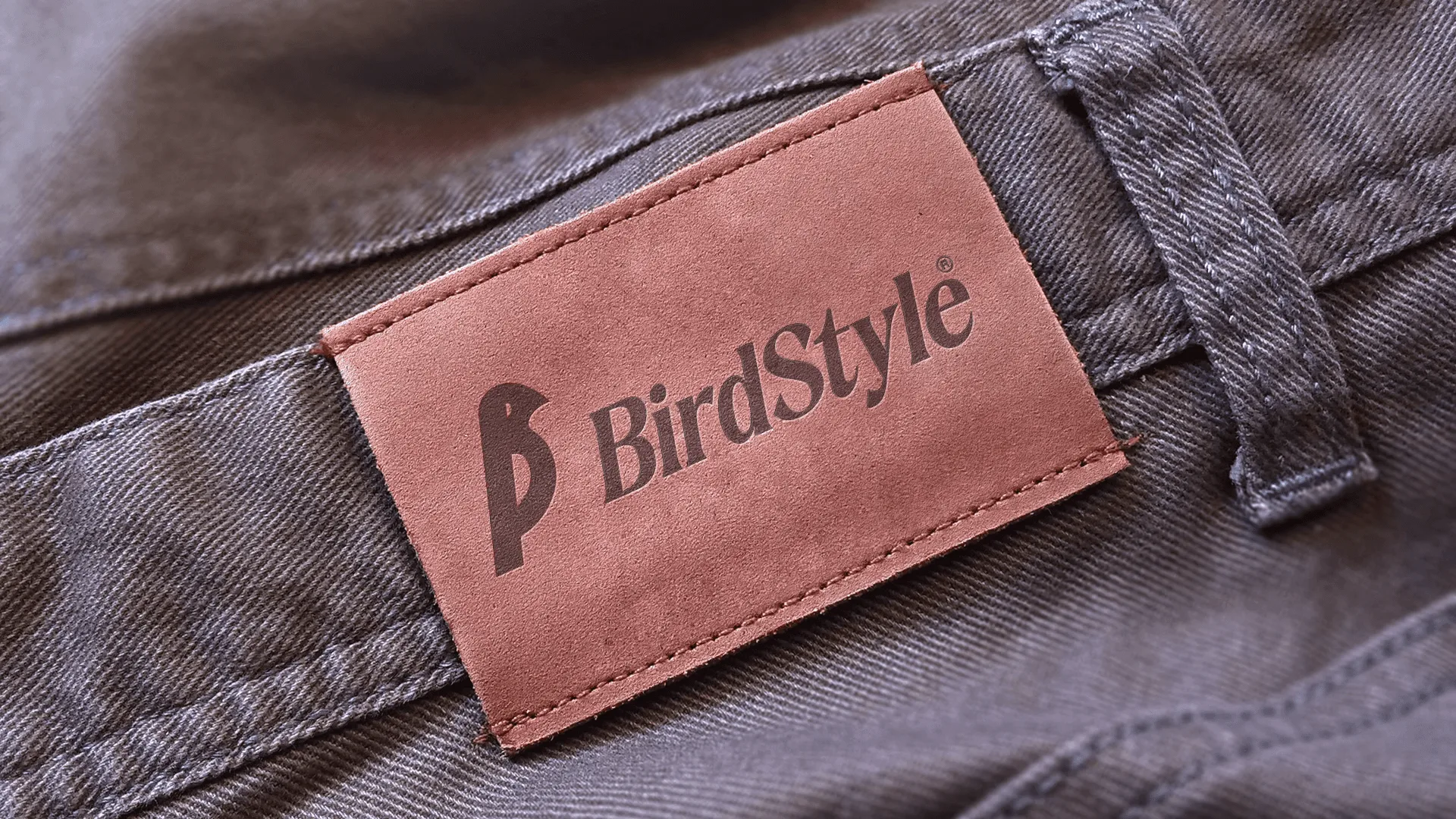 Etiqueta de couro rosada costurada no cós de uma calça cinza, com o símbolo e o logotipo BirdStyle