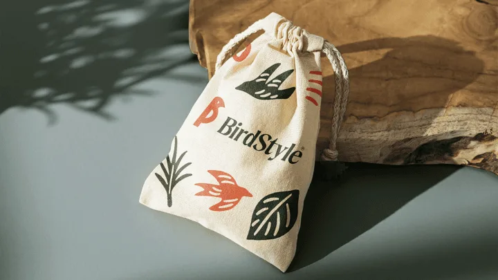 Saco de algodão cru com cordão, impresso com o logotipo BirdStyle e grafismos de folhas e pássaros em verde e laranja