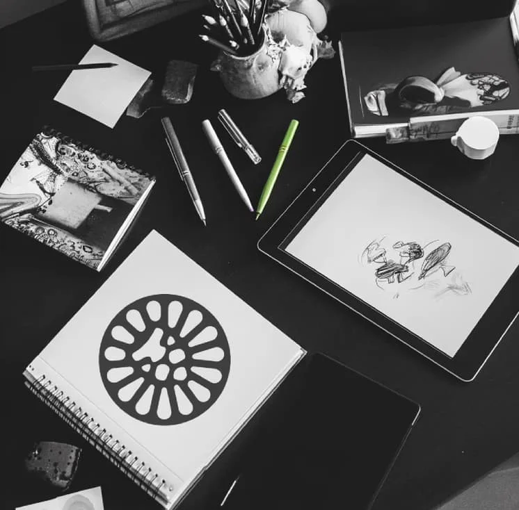 Mesa de trabalho em preto e branco com cadernos, canetas e um tablet exibindo esboços de design